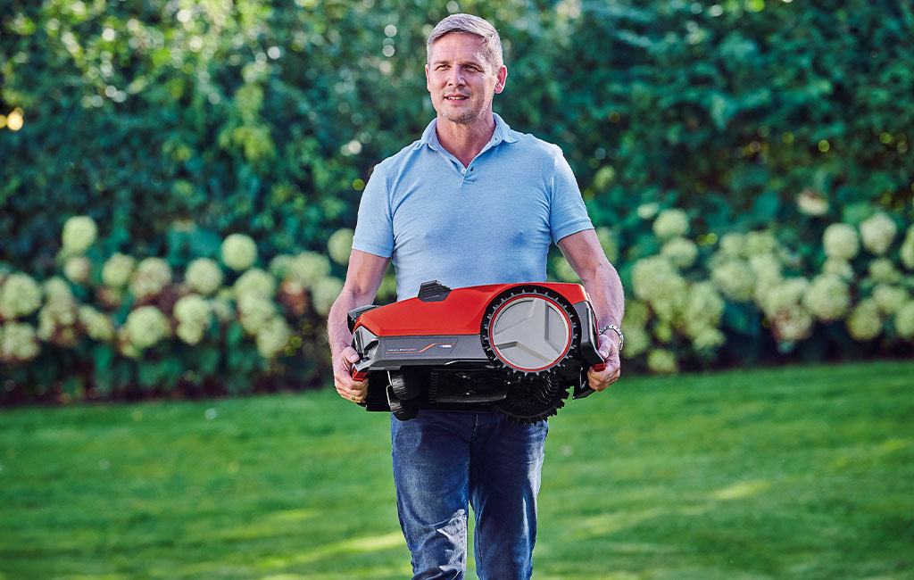 A man carrying a red robotic mower through the garden.