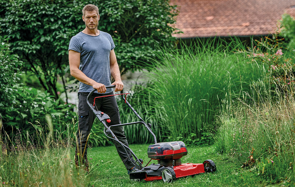 A man with a cordless mulching mower stand on a mowed strip of lawn surrounded by tall grown grass.