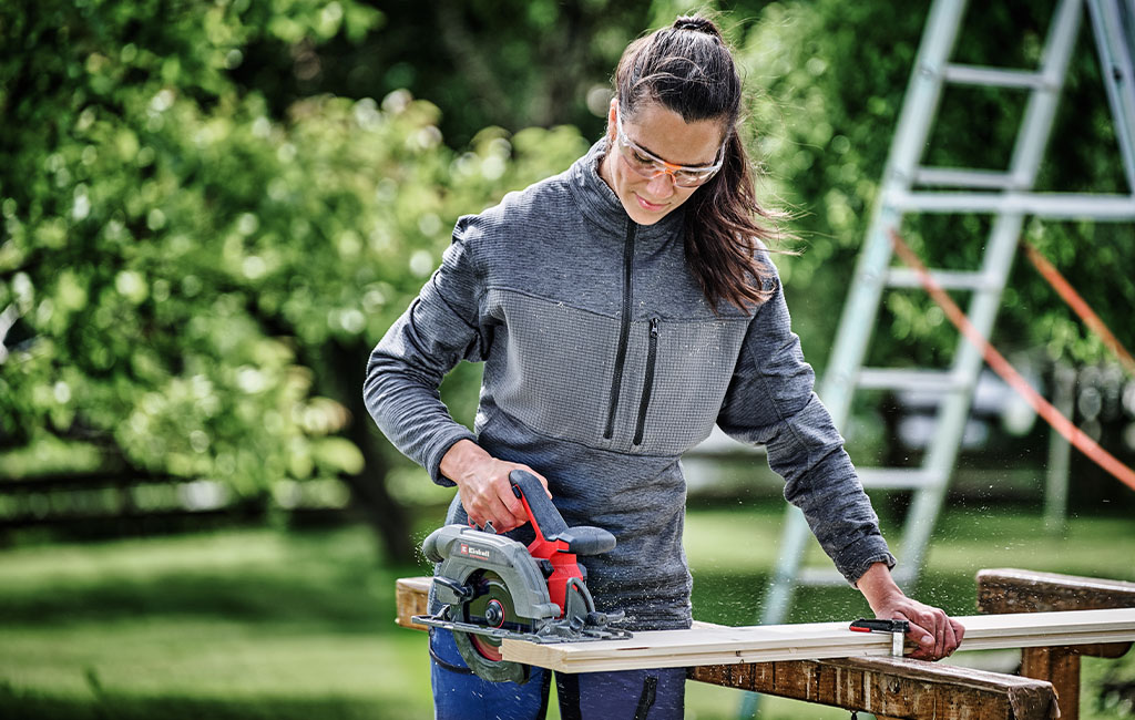 A woman wearing safety glasses is using a handheld circular saw to cut wood outdoors.