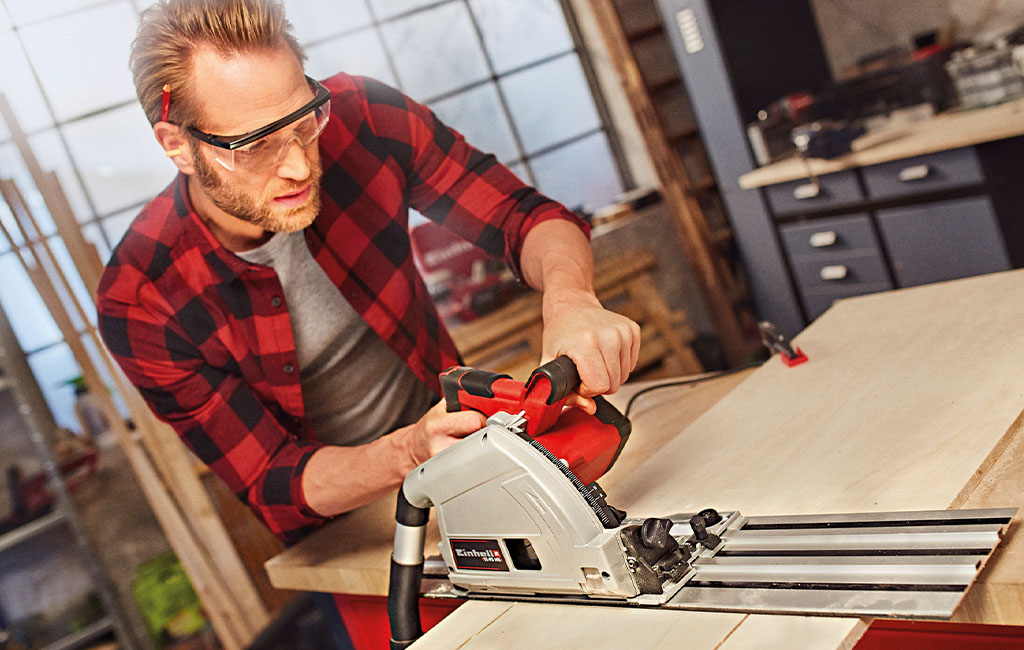 A man wearing safety glasses and a plaid shirt is working with a handheld circular saw in a workshop.