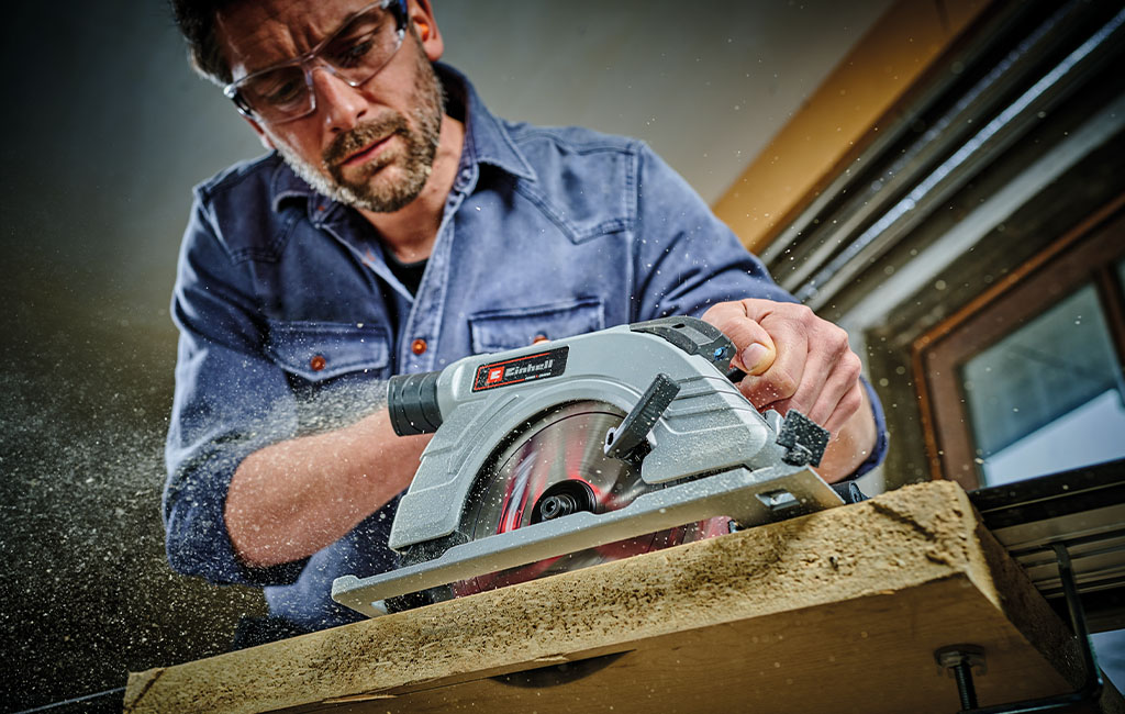 A man working with a handheld circular saw and wearing safety glasses on a piece of wood.