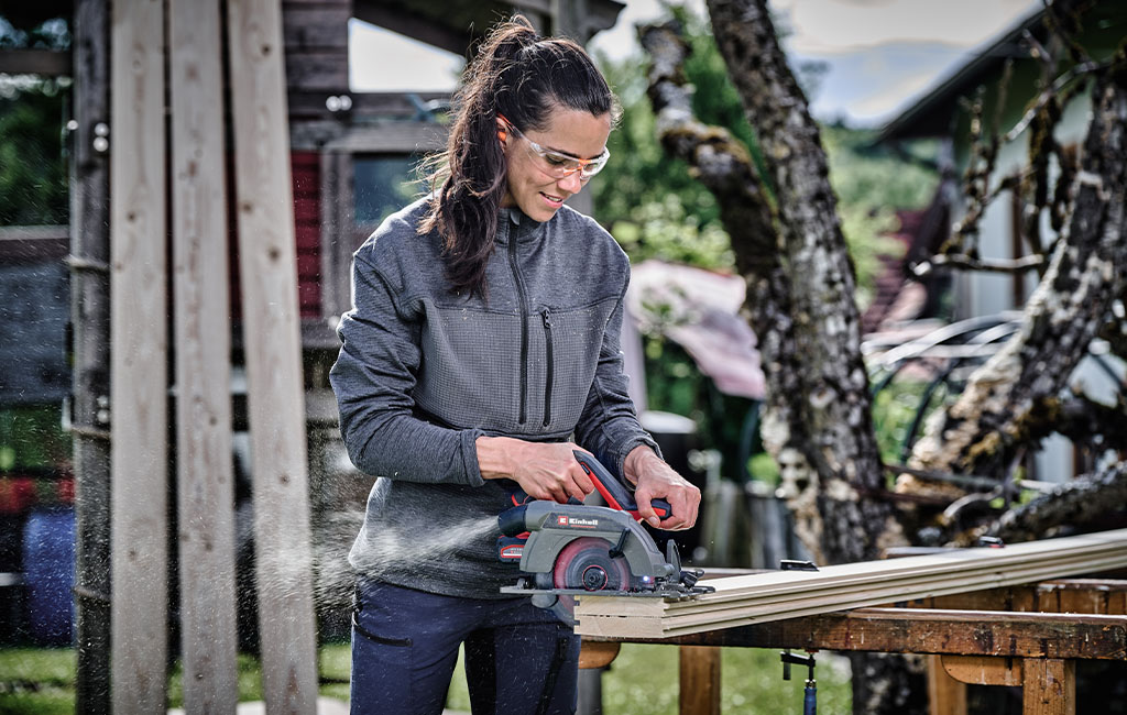 A woman wearing safety glasses is cutting wood with a handheld circular saw outdoors.