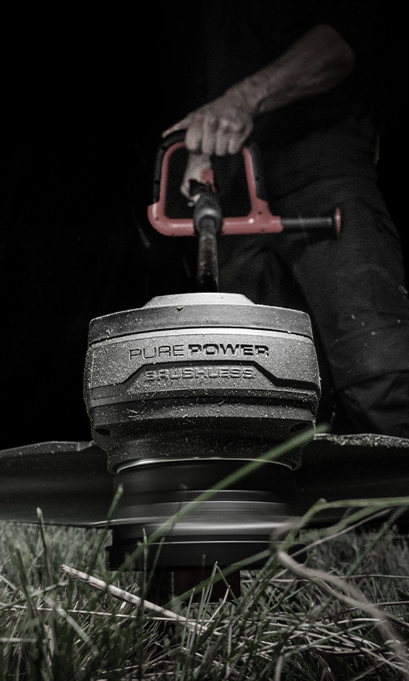 A red Einhell cordless grass trimmer is operated by a person in the background, with the focus on the spinning cutting head.