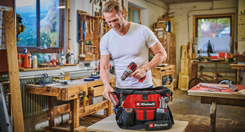 A tool bag, in the background a man with a cordless screwdriver in a workshop